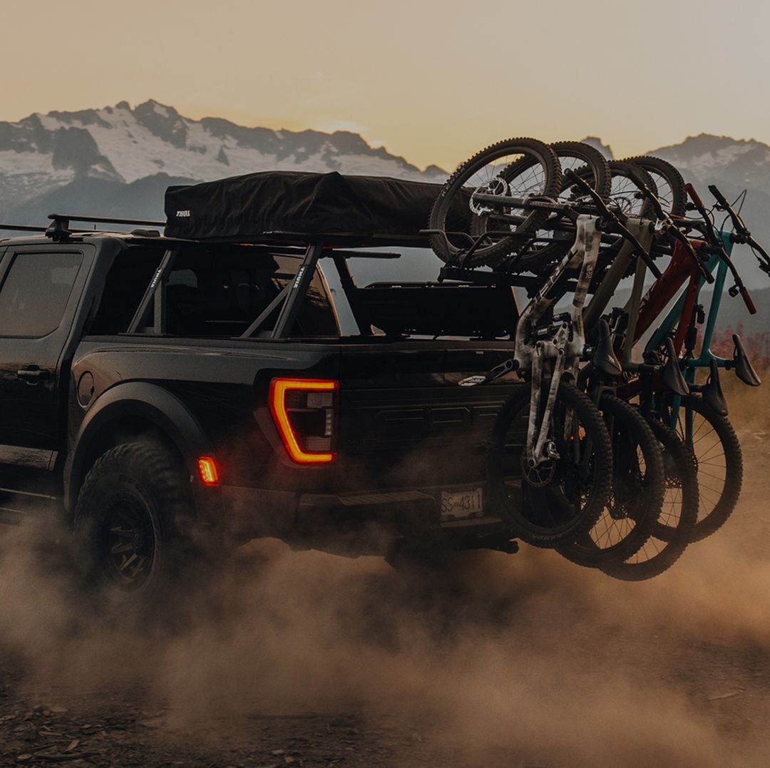 Camionnette roulant sur une route de montagne poussiéreuse au coucher du soleil avec plusieurs vélos montés sur un porte-vélos Thule Revert, faisant la promotion de « Amenez votre vélo ».