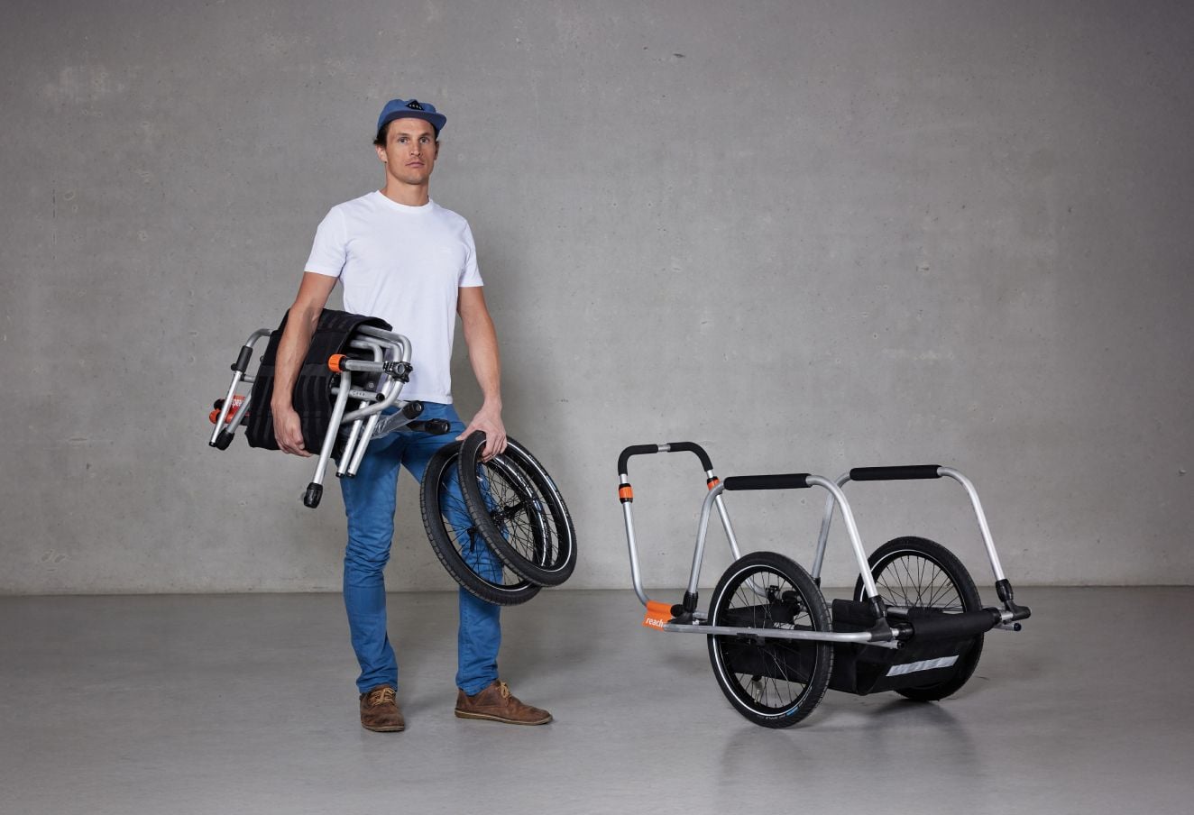 En man står i en studio med en hopfälld Reacha Sport cykelvagn och visar upp dess lätta och kompakta design.