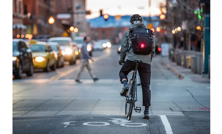 A bike commuter waits at a street crossing, wearing a Thule Pack 'n Pedal backpack with a red bike light attached.