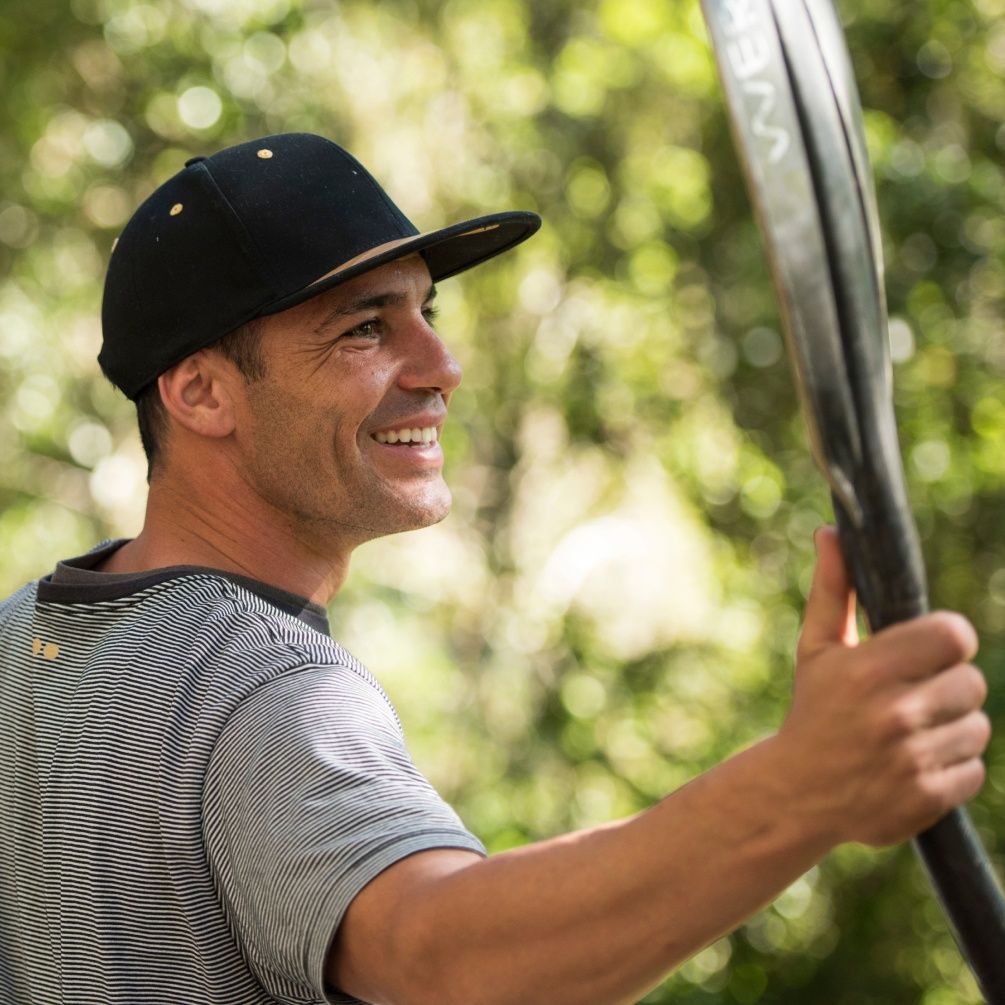 A close up image of Pedro Oliva wearing a black baseball hat and smiling while holding a kayak paddle.