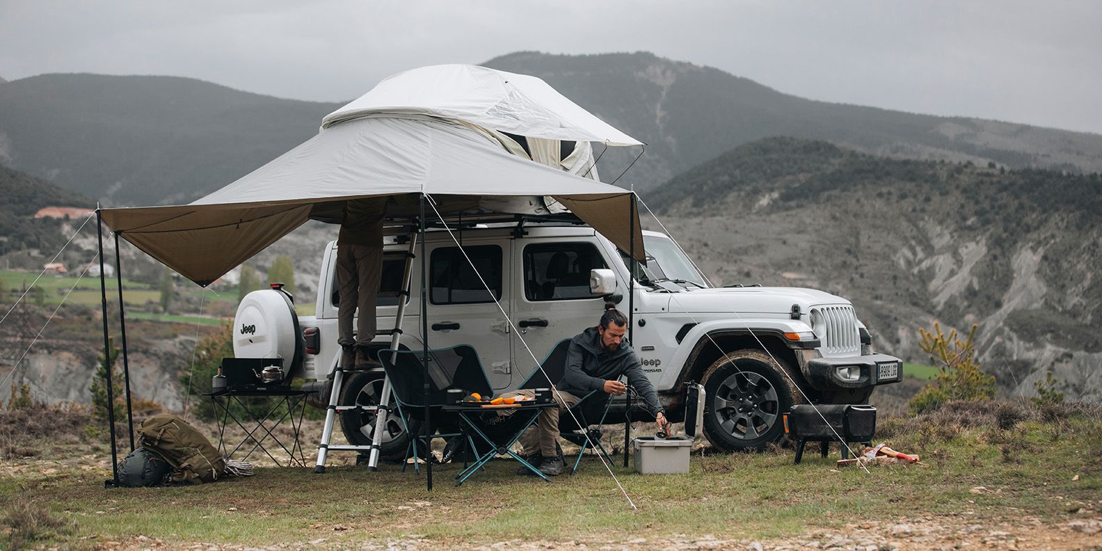 In the mountains a jeep is parked with a Thule Approch rooftop tent and a man sits under the awning
