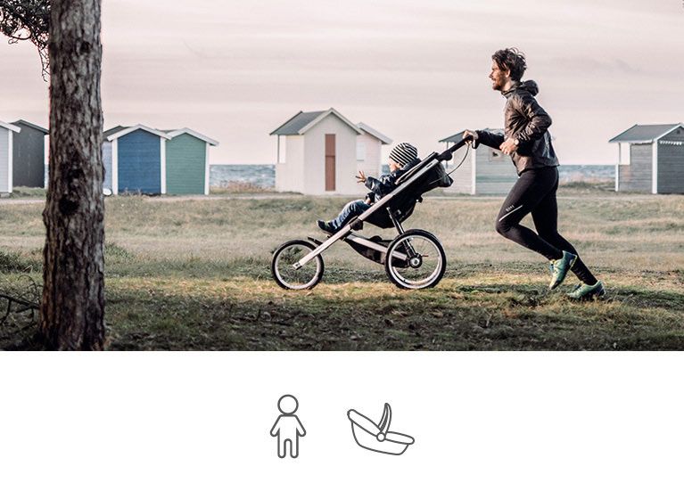 A father jogs with his child in a thule baby stroller on the grass by small houses on the beach.