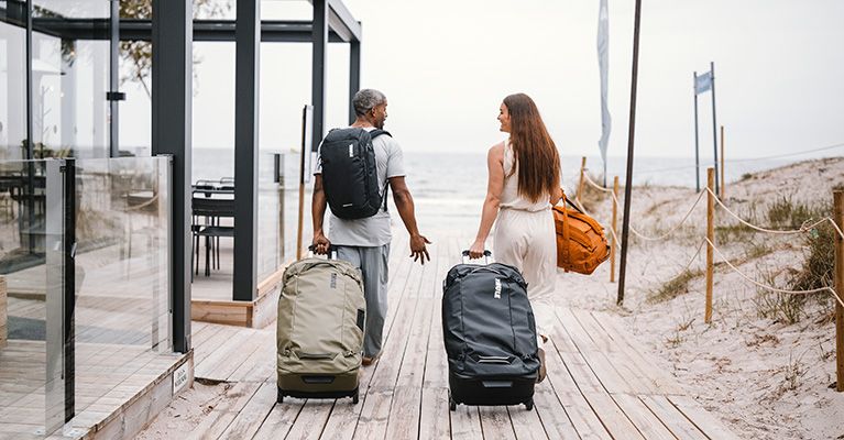 A man and woman walk down boardwalk of a beach with Thule suitcases and backpacks