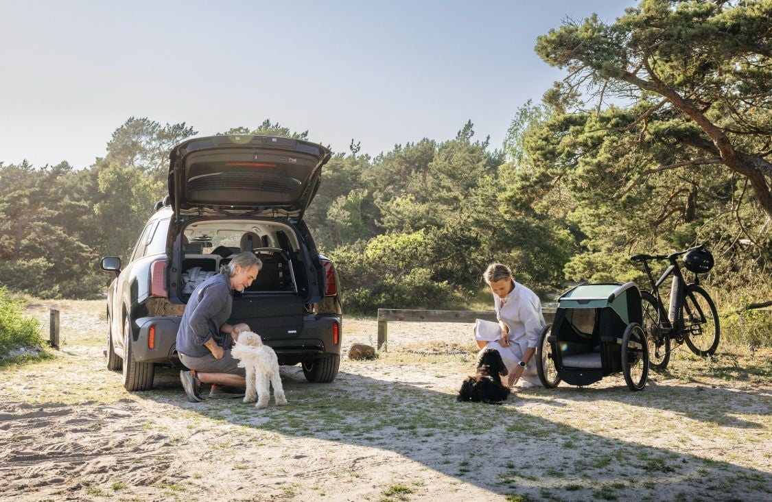 Um homem faz festas a um cão atrás de um automóvel com um transportador para cães Thule Allax, enquanto uma mulher faz festas a um cão junto a um atrelado para bicicleta Thule Bexey. 