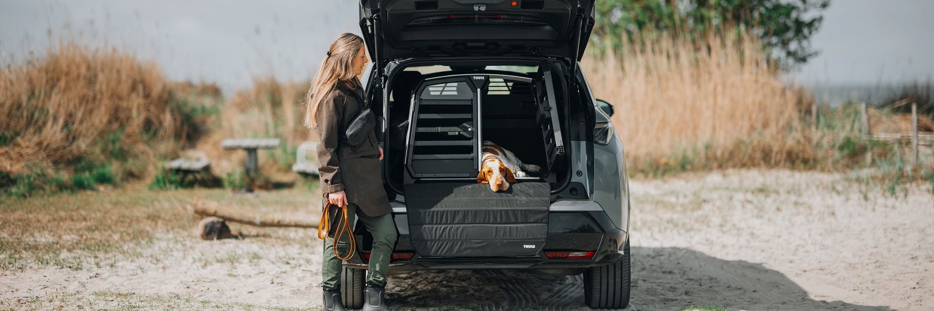 A woman stands beside an SUV with the rear hatch open, where a dog relaxes inside a Thule Allax dog crate. The scene highlights safe and comfortable travel for dogs in an outdoor environment.