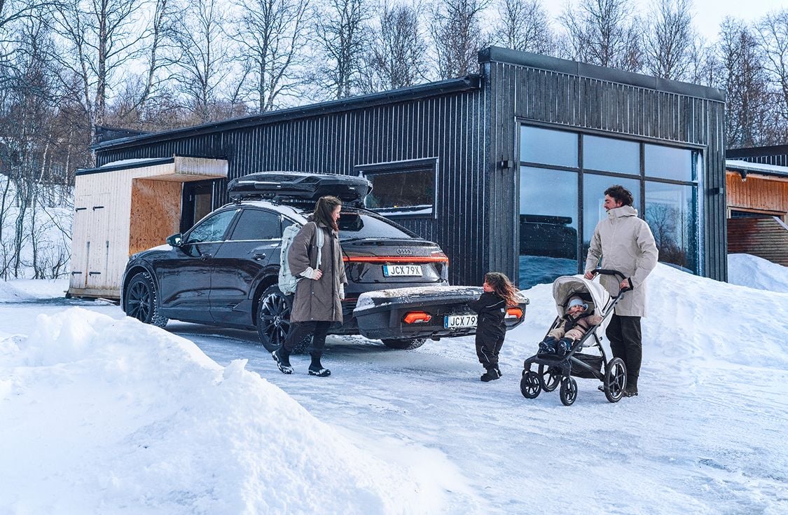 Obitelj stoji pokraj tamnog SUV-a s kutijom za križanje i teretnim nosačem Thule Arcos smještenim straga. Parkiran je ispred moderne kolibe u snježnom krajoliku, s jednim odraslim koji gura kolica, a dijete ide prema vozilu.