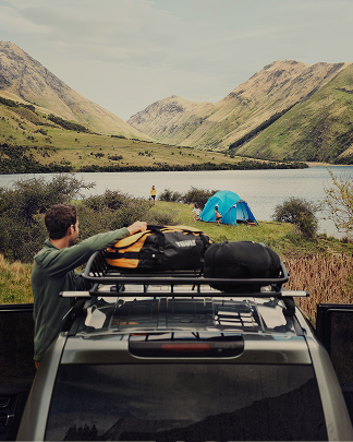 Um homem carrega uma bolsa de viagem Thule em uma plataforma de teto Thule Caprock, no teto de um veículo, com barracas e um lago de montanha ao fundo.