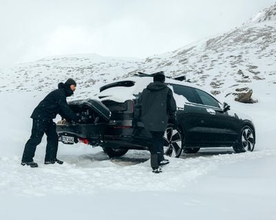 Two people standing behind a car,  loading gear into an open rear‑mounted cargo carrier.