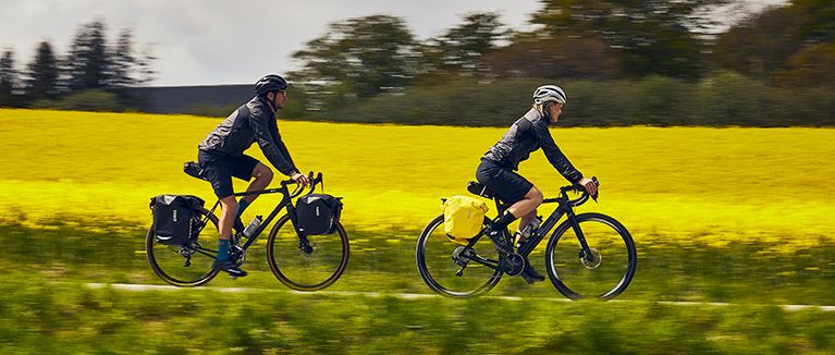 Two cyclists bike past a yellow field with black and yellow bike panniers.