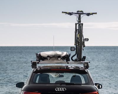 A car is parked by the water with a surfboard and bike on the roof racks.