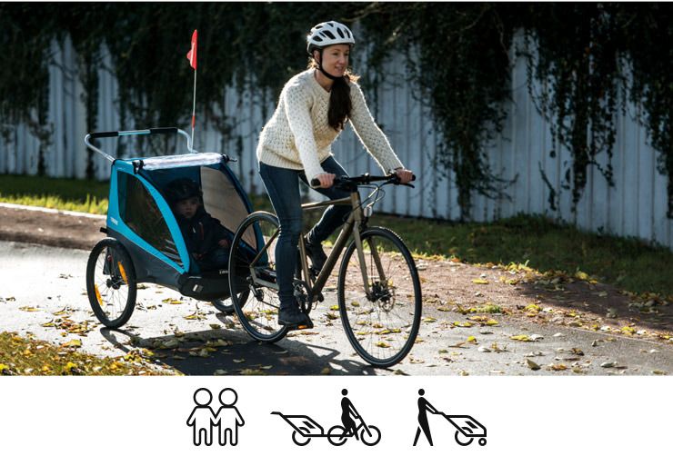 A woman bikes down a bike path with her child in Thule Coaster XT bike trailer for kids.