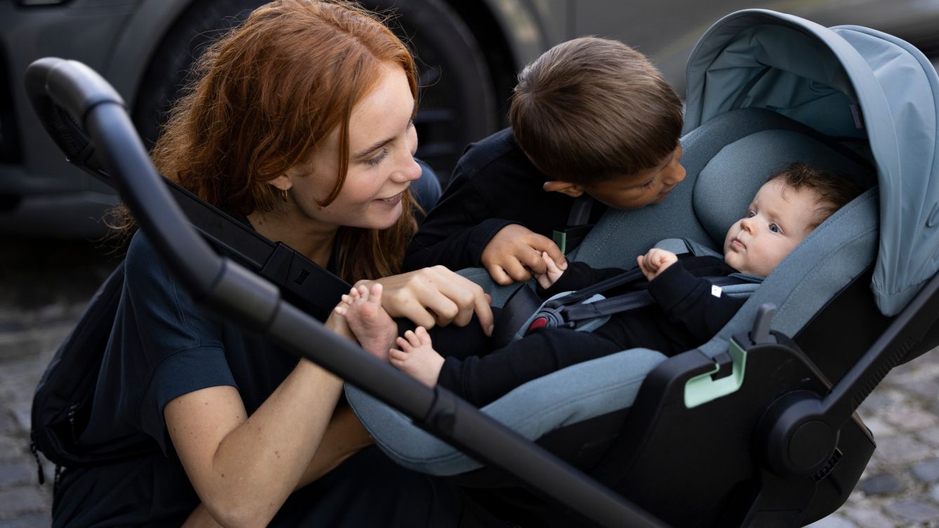 Una mujer se agacha para observar un asiento de niños para automóvil Thule Maple montado en una carriola, con un bebé en su interior y un niño pequeño de pie a su lado.