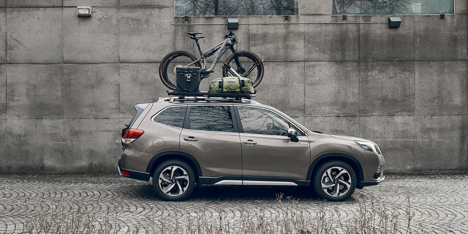 A vehicle parked next to a wall with a Thule Caprock roof platform, bike, duffel and other gear on the roof.