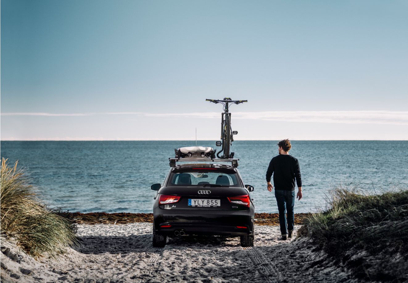 En person går ved siden av en bil som har en sykkel på taket og er parkert på en strand.