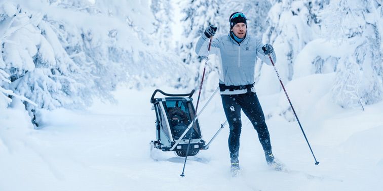 A man goes cross-country skiing in the woods with a blue Thule Chariot cross-country bike trailer.