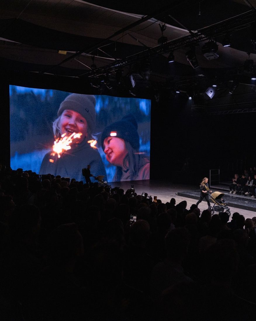 A performer on a runway in front of a large screen displaying a close-up winter scene, with the audience watching.