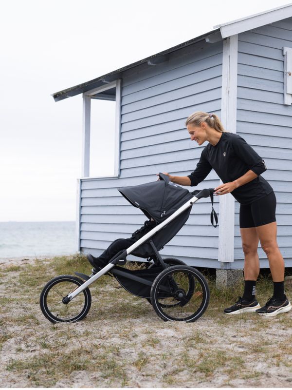 海边小路上，一名穿着跑步装备的女性在海边小屋旁调整 Thule Glide 3 轻便婴儿车