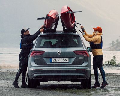 Два человека выгружают каяк из припаркованного на берегу автомобиля с креплениями для водного спорта.