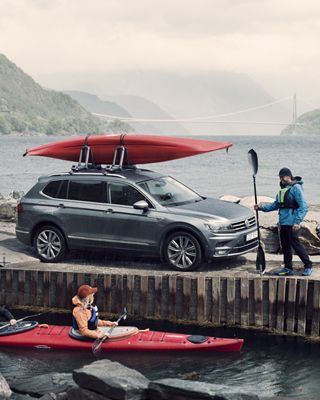 A man stands at the dock next to a car with a kayak rack, and a woman in a kayak in the water talks to him.