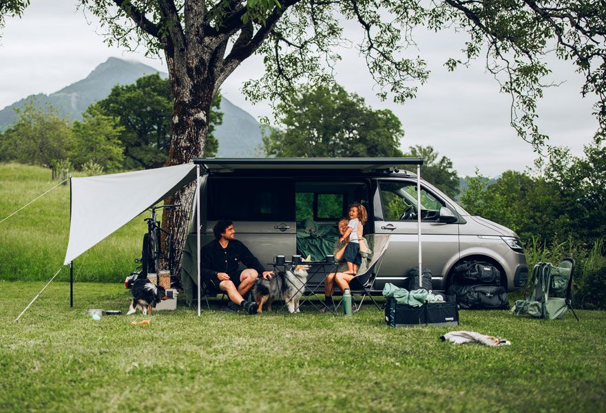A compact van is parked in the grass with a Thule Subsola awning panel and a family sits underneath.