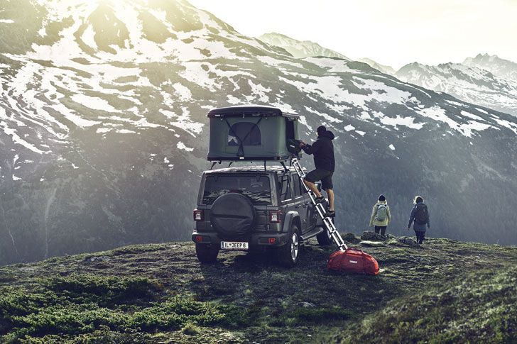 A man climbs into a hard-shell rooftop tents on a jeep parked next to a snowy mountain.