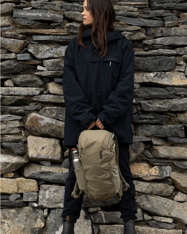 Eine Frau lehnt an einer Steinmauer und hält einen Thule Rucksack.