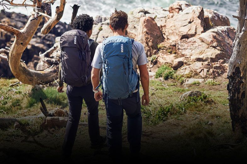 Two people with Thule hiking backpacks walk along a rocky coast.