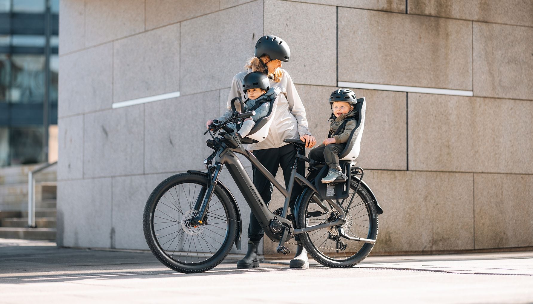 Adult standing with a bicycle fitted with Thule Yepp Nexxt 2 mini and Thule Yepp Nexxt 2 maxi child seats, each carrying a child, beside a concrete building wall.