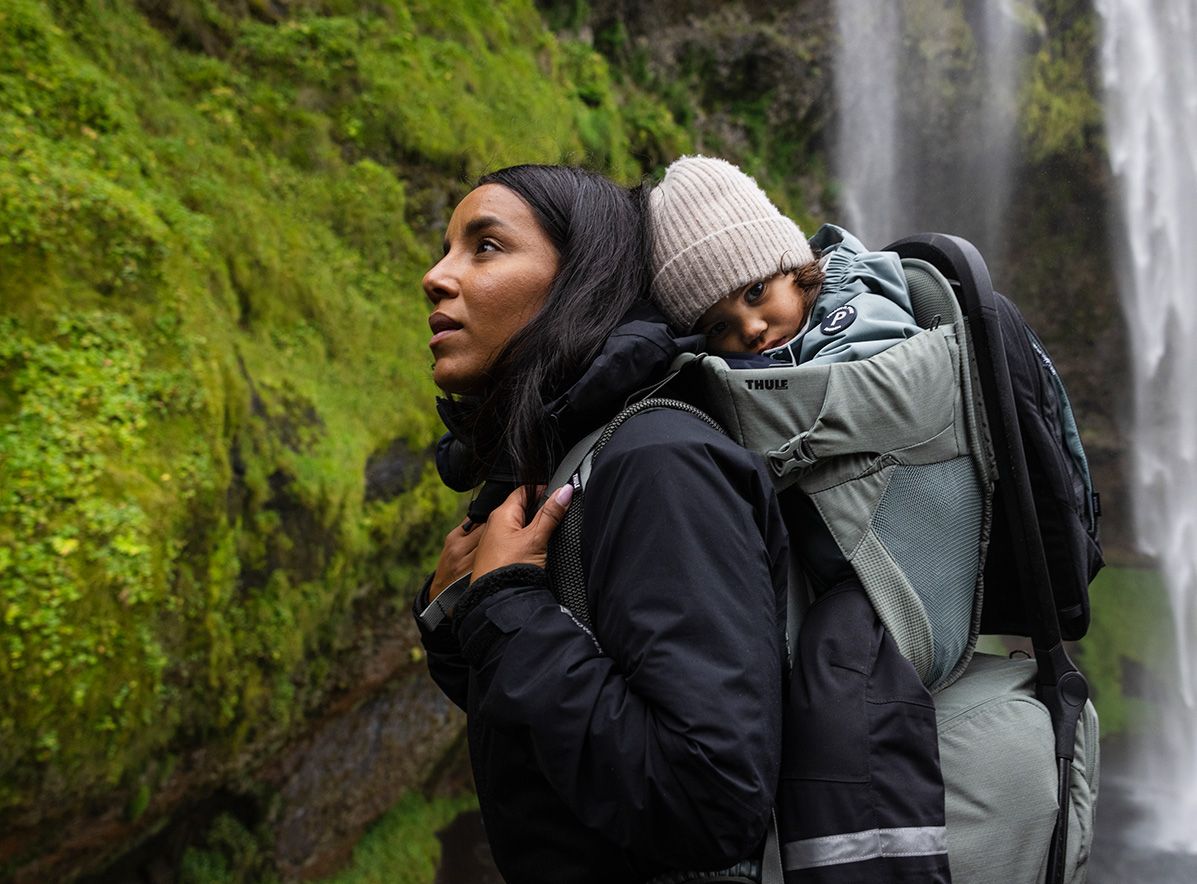 A woman carries her baby in a child carrier backpack next to a waterfall.