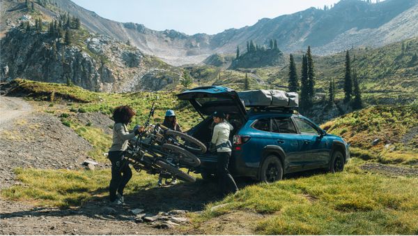 Three cyclists load their bikes off the bike rack on a vehicle with the trunk open parked in the mountains.