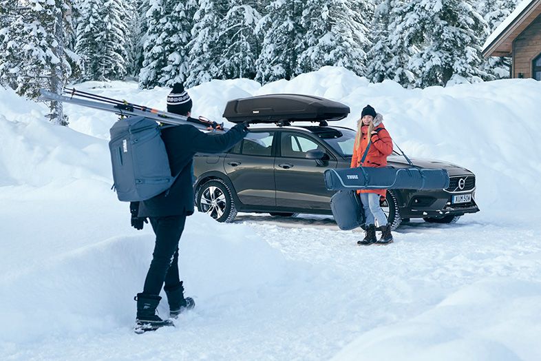 A man walks through the snow with a ski boot bag towards a woman by a car with a ski travel bag and backpack.