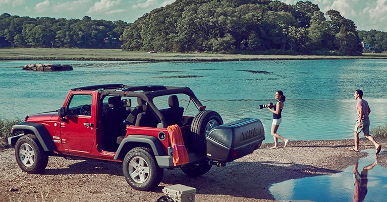 A jeep is parked beside a lake with a Thule Combi hitch cargo carrier and two people taking photos.