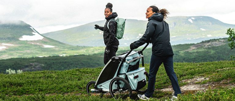 Two women walk through a grassy, hilly landscape with a light blue Thule Chariot multisport bike trailer.