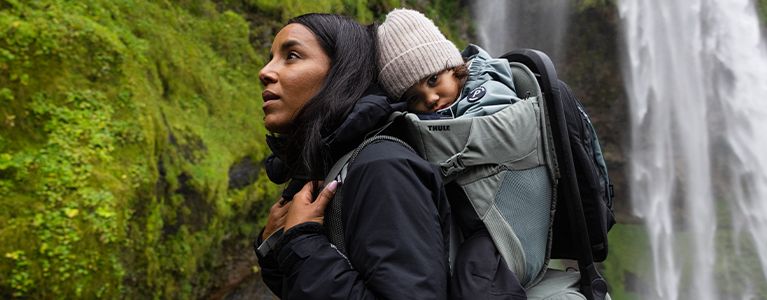 By a waterfall, a woman looks up with a baby in one of the Thule baby carrier backpacks.