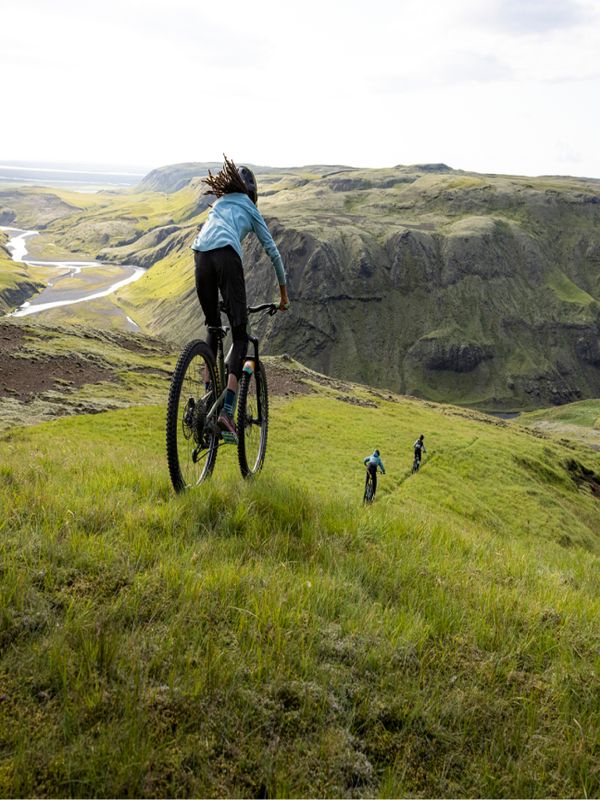 Un ciclista segue altri due ciclisti in lontananza giù per una collina verde di gra con cielo azzurro e un fiume.