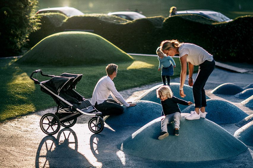 In a park with interesting bumpy shapes, parents play with their kids next to a Thule twin stroller.