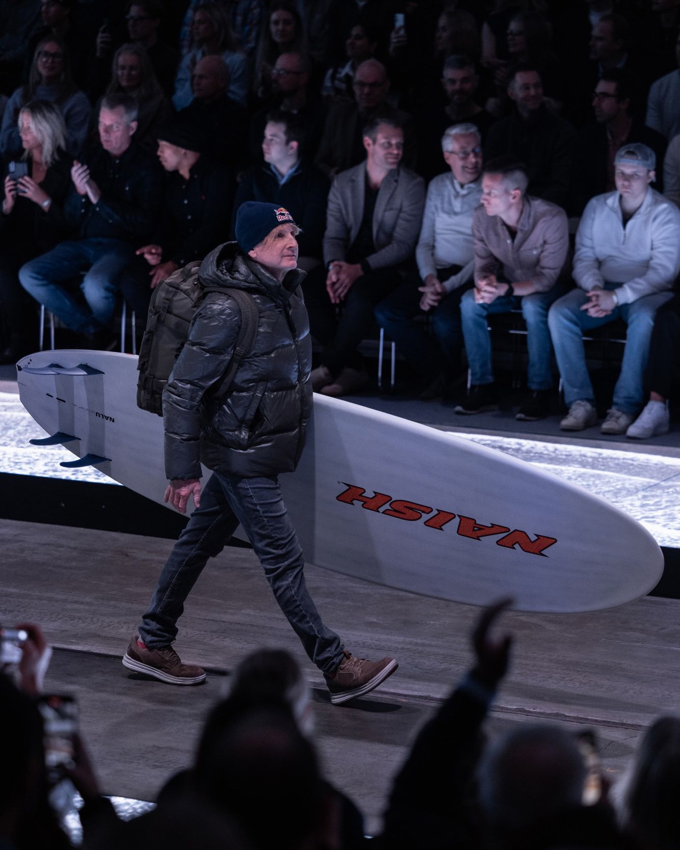 Robby Naish walking down a runway carrying a large surfboard, with an audience seated on both sides.