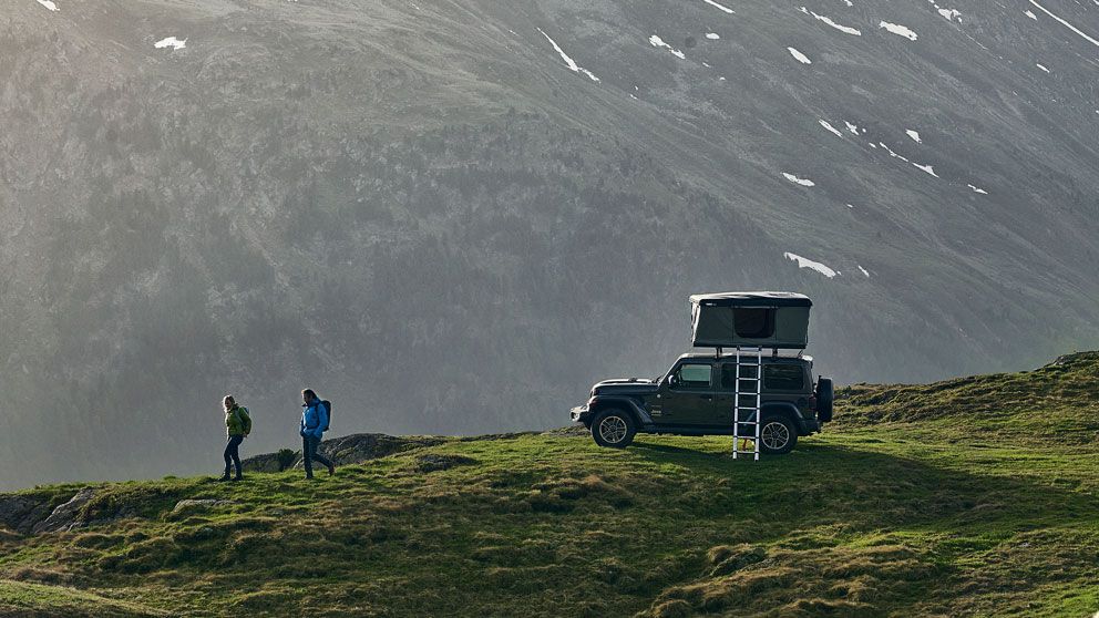 Two people hike along a grassy path next to a vehicle with a hard shell roof top tent of the roof racks.