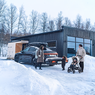 Una famiglia cammina vicino al retro di un'auto montata con una scatola sul tetto Thule sopra e un trasportatore cargo posteriore Thule sul retro in un vialetto innevato.