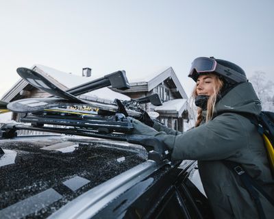 Eine Frau, die eine Skibrille trägt, lädt ihre Ski mithilfe von Winterträgerzubehör auf das Dach ihres Autos.