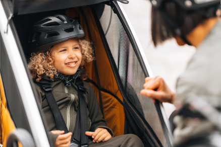 A child sits smiling inside a bike trailer while an adult leans beside it.