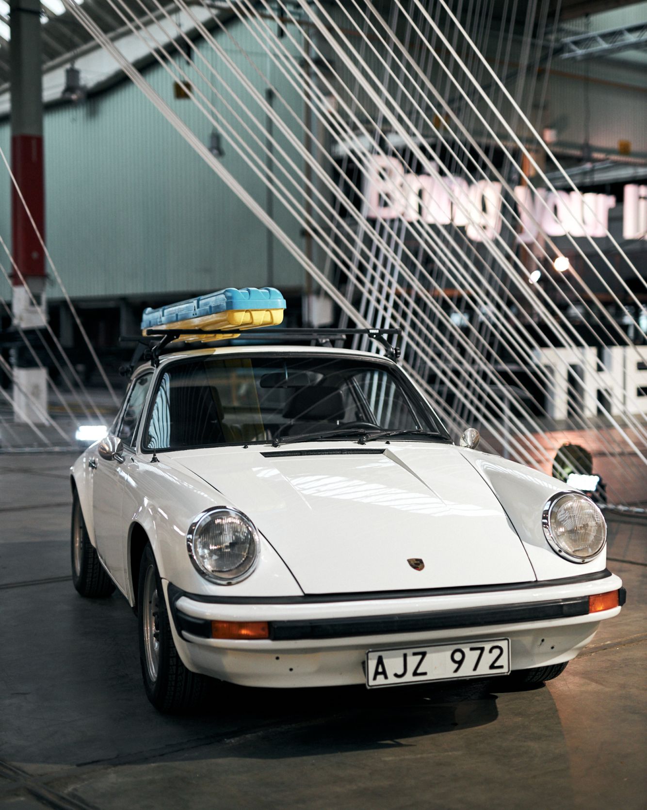 A white vintage Porsche 911 is parked in an exhibition hall with a vintage Thule roof box mounted