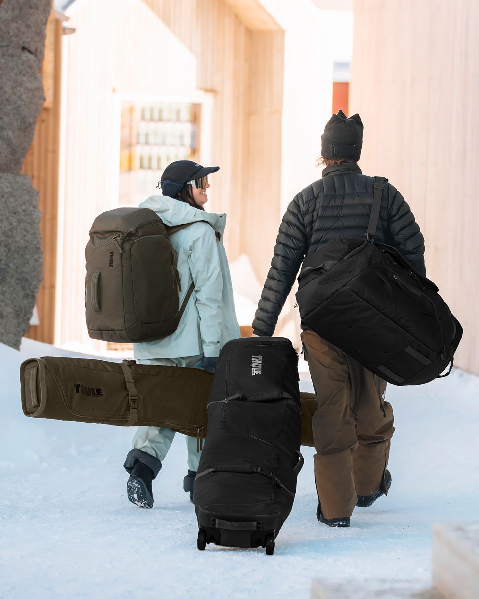 Duas pessoas a caminhar, carregando uma mochila lage, um grande saco de viagem, um saco de esqui e a enrolar uma mala de viagem com rodas.
