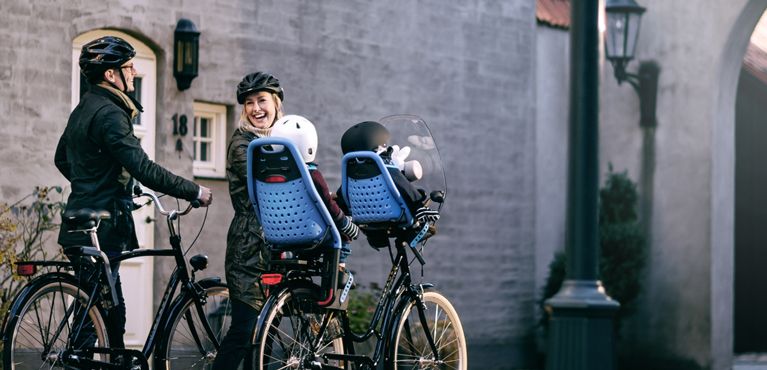 A woman and man laugh, holding bikes, one with kids in the kids bike seat and a baby in the baby bike seat.