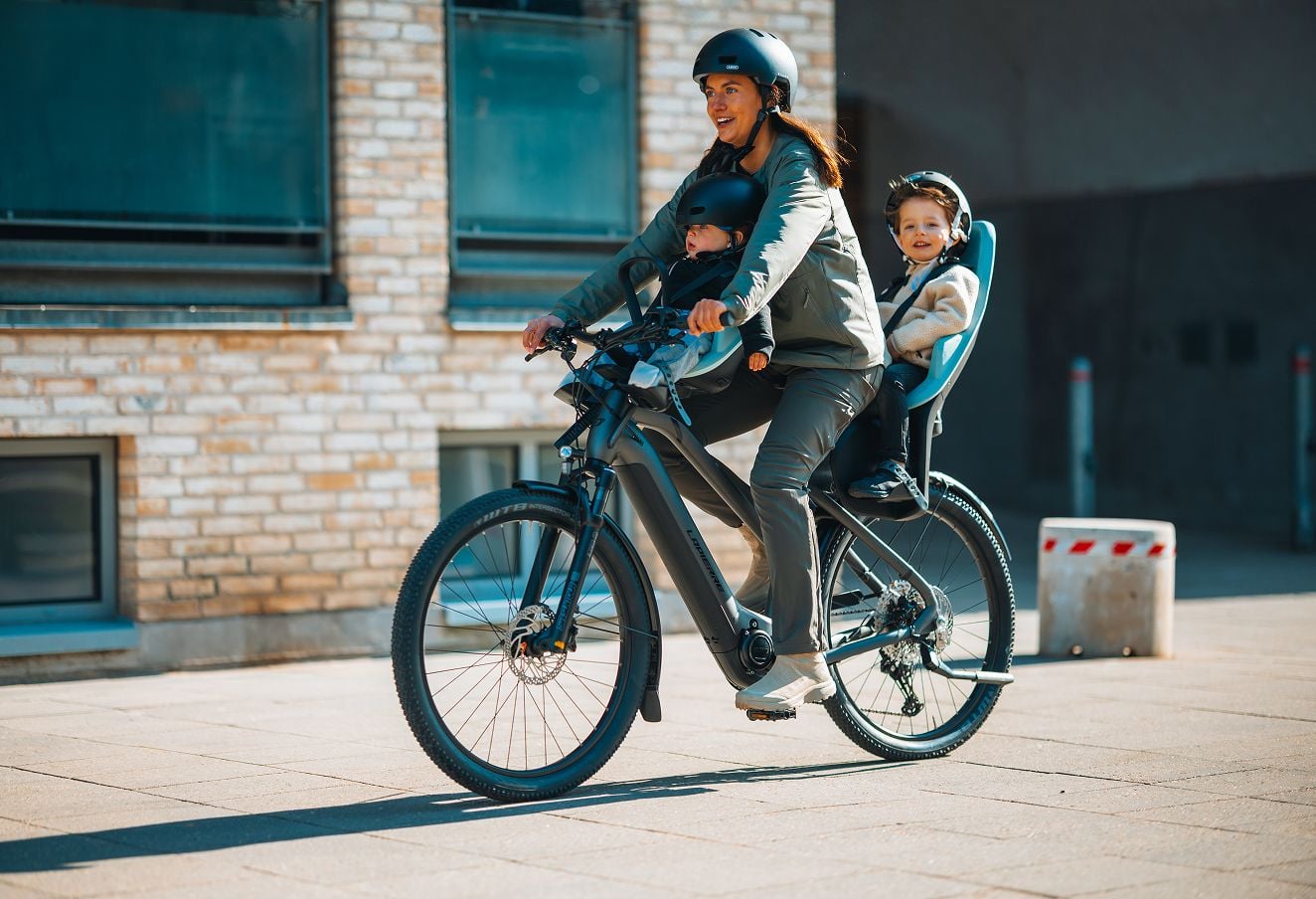 Person riding an e-bike with two children seated in Thule Yepp 2 front and rear child seats along a city sidewalk beside a brick building.