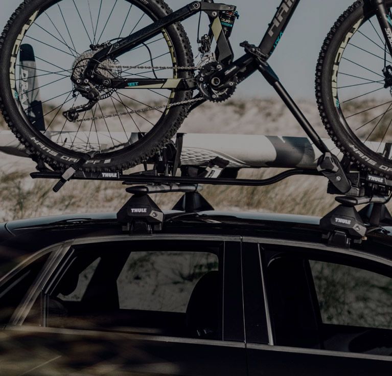 On a sandy beach a man walks beside a car with Thule Evo Wingbar roof racks with a bicycle and SUP board attached.