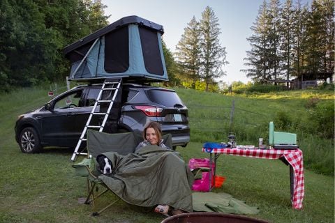 Woman  with a dog tucked beside her in a camp chair and an SUV with an open rooftop tent behind