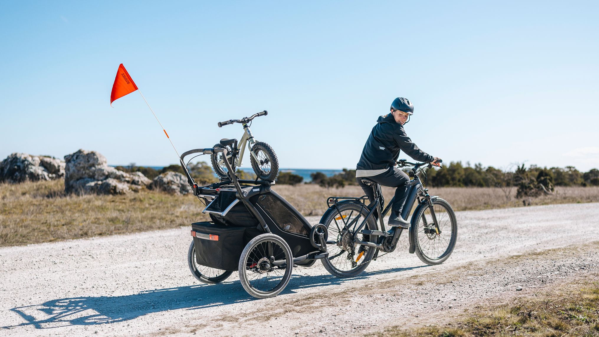 Charlotte Kalla pedalando por uma estrada de cascalho, rebocando um trailer de bicicleta Thule com uma bicicleta infantil montada no topo, por uma paisagem costeira aberta sob um céu azul claro.