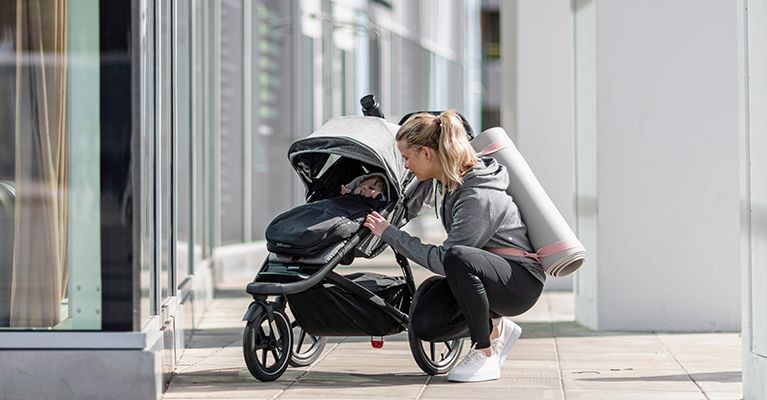 A woman with a yoga mat kneels down by a stroller to tend to her baby in a Thule footmuff.