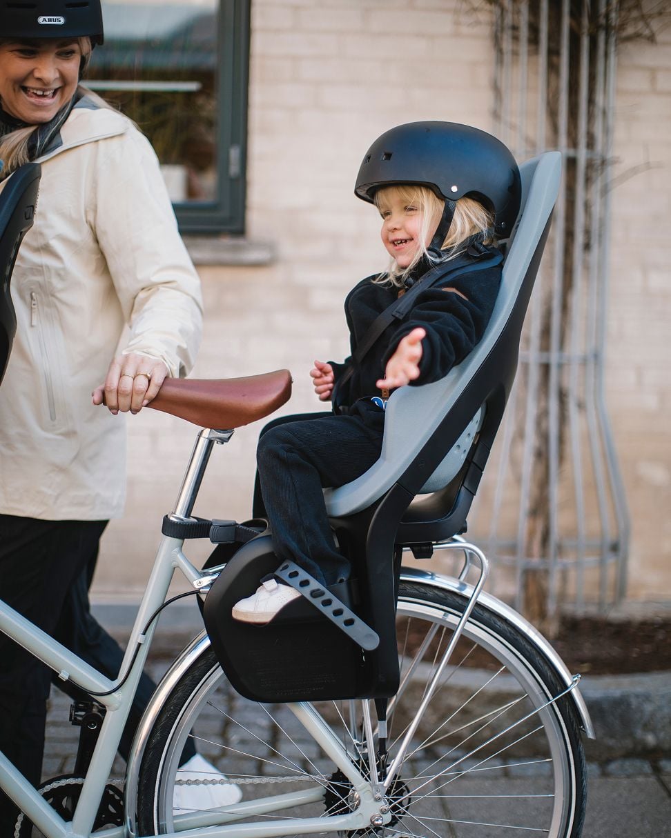 Child seated in a rear bike seat reaching out while an adult stands beside, holding the bicycle, in front of a brick building.